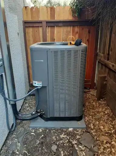 Gray HVAC condenser unit installed on concrete pad against home exterior with wooden fence