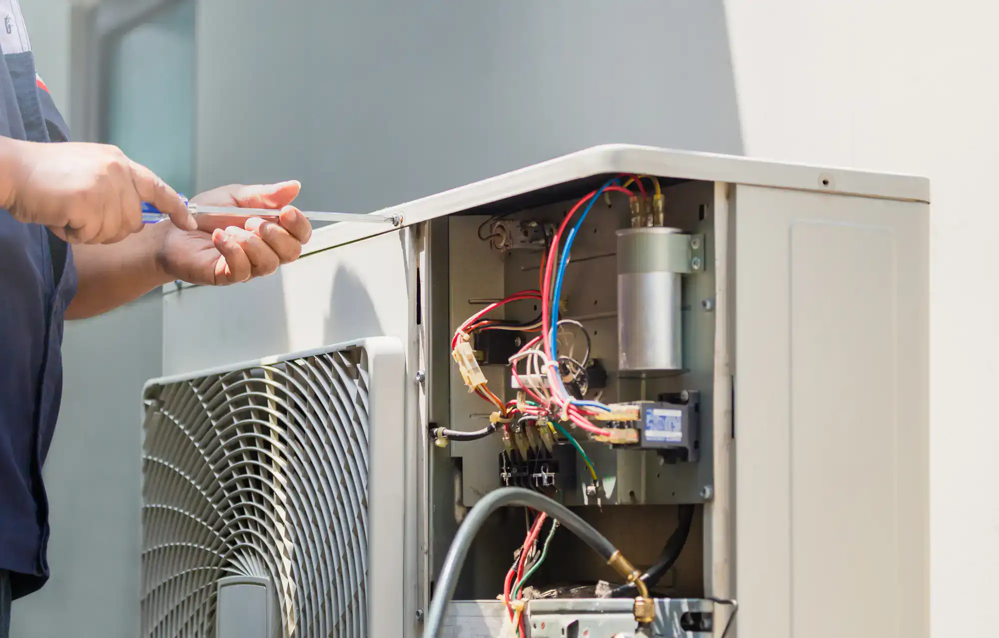 A person uses a screwdriver to repair or service the electrical components inside an open outdoor air conditioning unit. Colorful wires and mechanical parts are visible inside the unit.