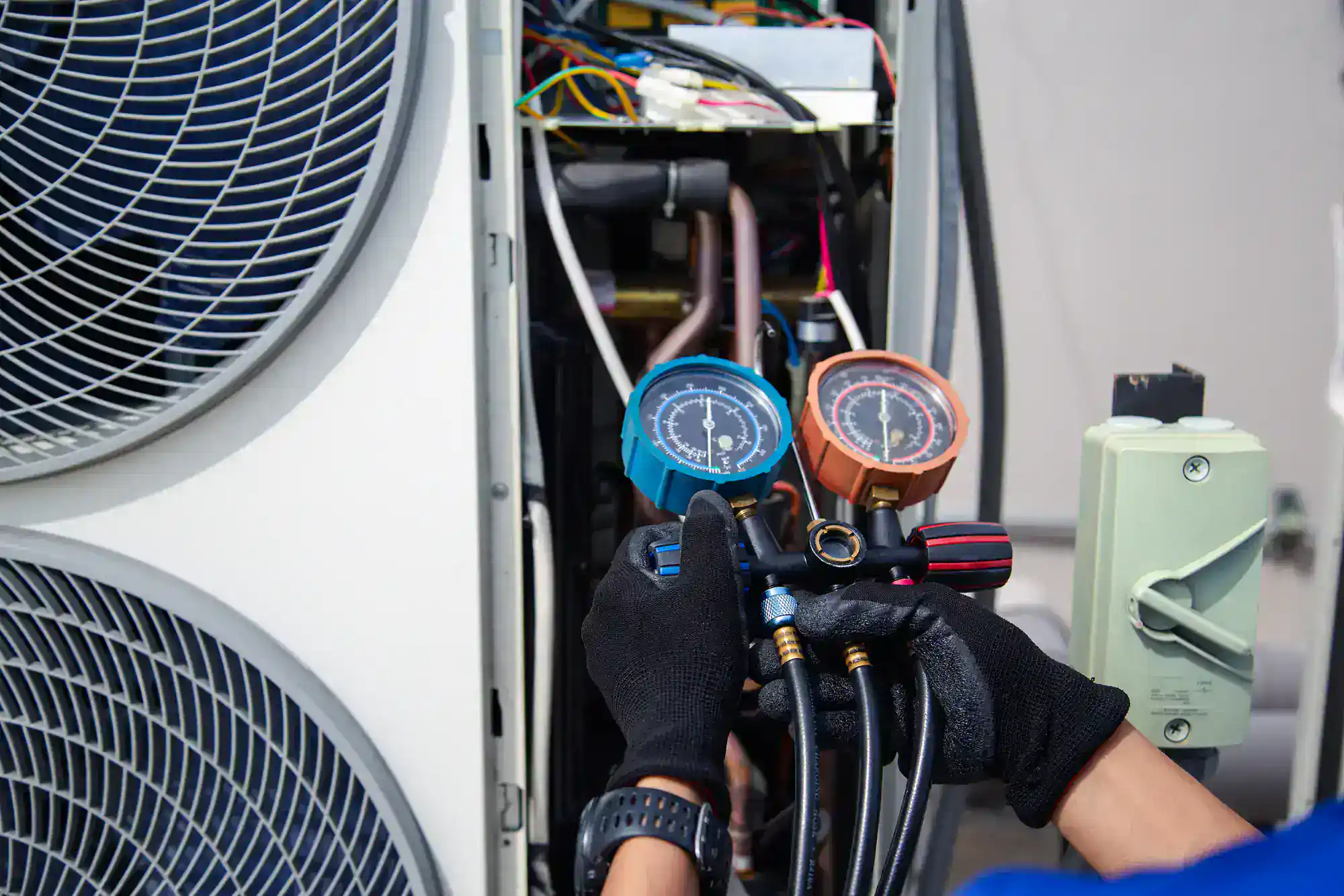 A technician wearing black gloves uses a pressure gauge manifold to check an air conditioning unit with exposed wiring and components.