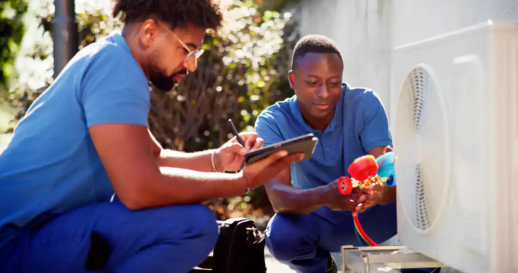 Two technicians in blue uniforms work on an outdoor air conditioning unit; one adjusts gauges on the unit while the other writes on a clipboard. They are focused and appear to be performing maintenance or repairs.