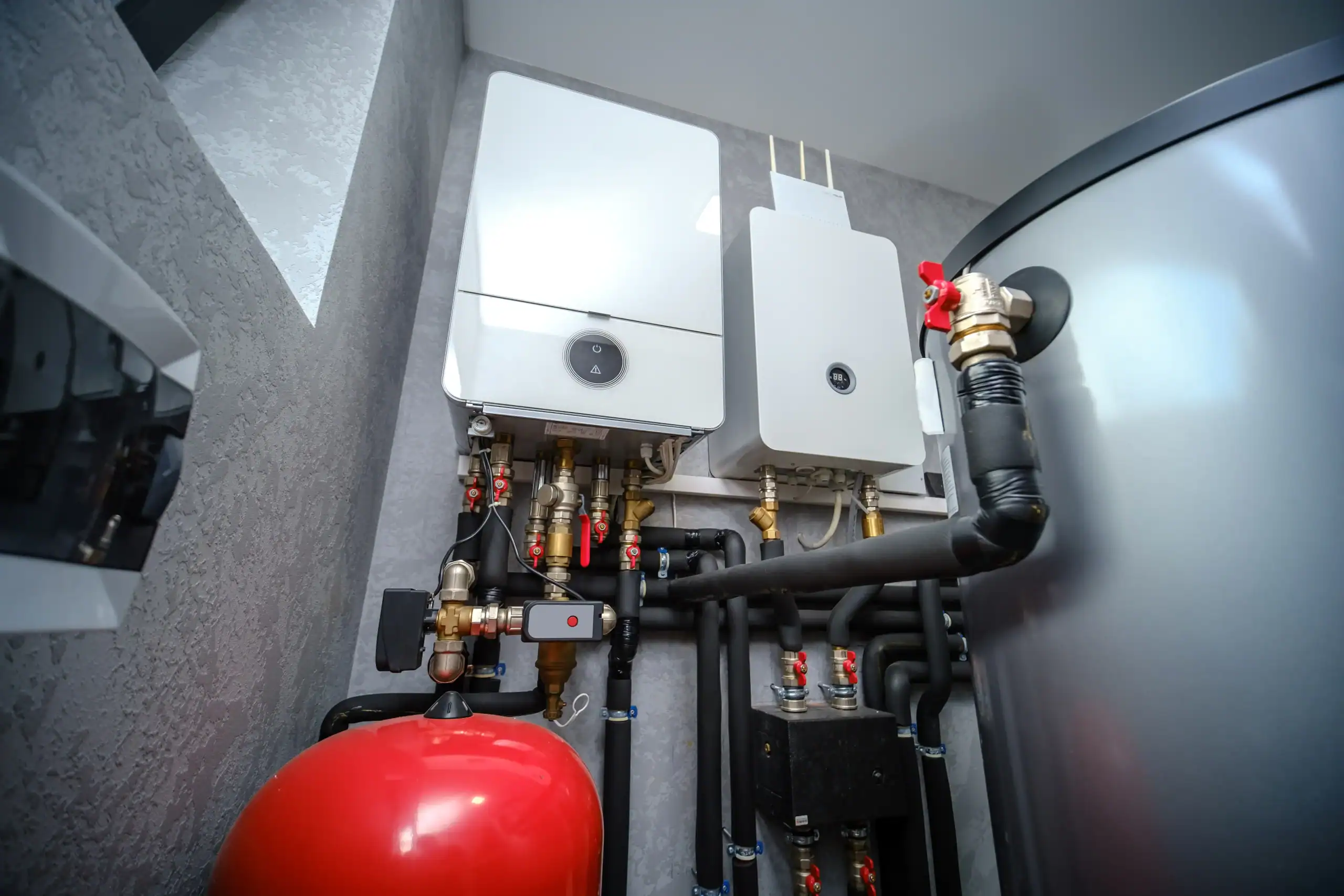 Modern boiler room with two white wall-mounted boilers, multiple pipes and valves, a large gray water tank, and a red expansion tank, all installed against a light-colored wall.