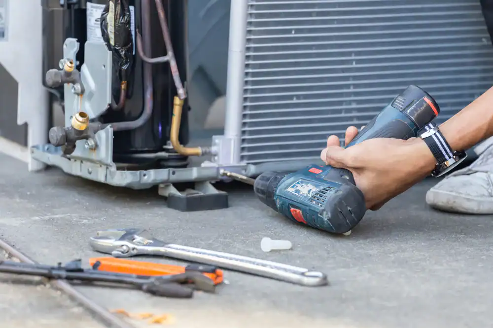 A person uses a cordless power drill to repair or install an air conditioning unit, with various hand tools like a wrench and pliers lying on the ground nearby.