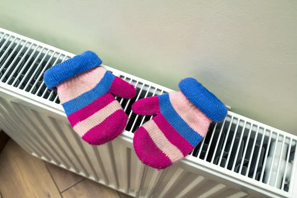 A pair of colorful striped mittens with blue cuffs, pink, purple, and blue stripes resting on top of a white radiator against a light-colored wall.