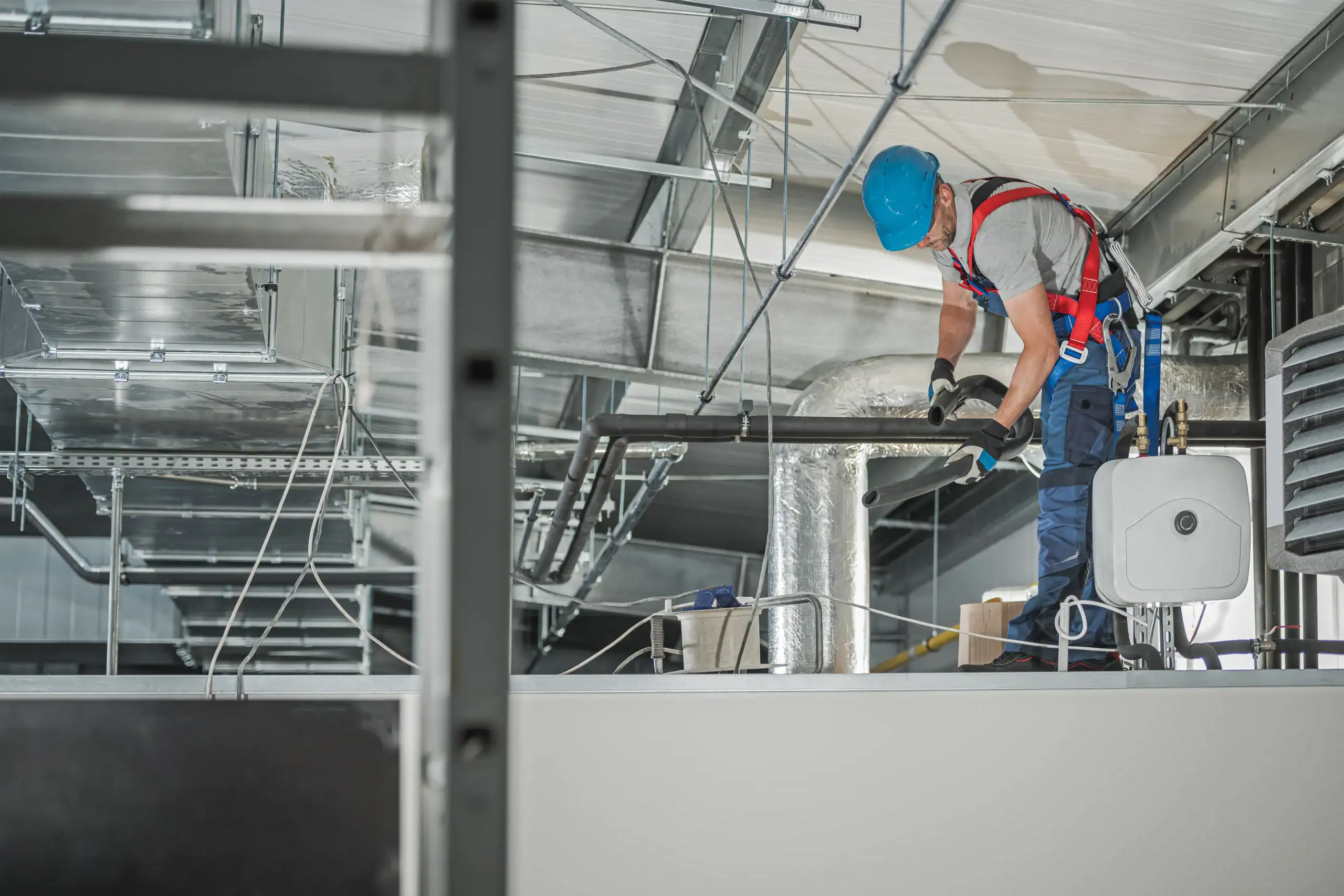 A worker wearing a blue hard hat and safety harness installs or repairs pipes on a raised platform inside an industrial building with ventilation ducts and cables overhead.