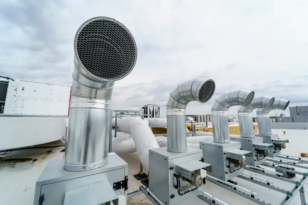 Four large, silver industrial air vents or exhaust ducts are installed on a building rooftop under a cloudy sky, with additional HVAC equipment and city buildings visible in the background.