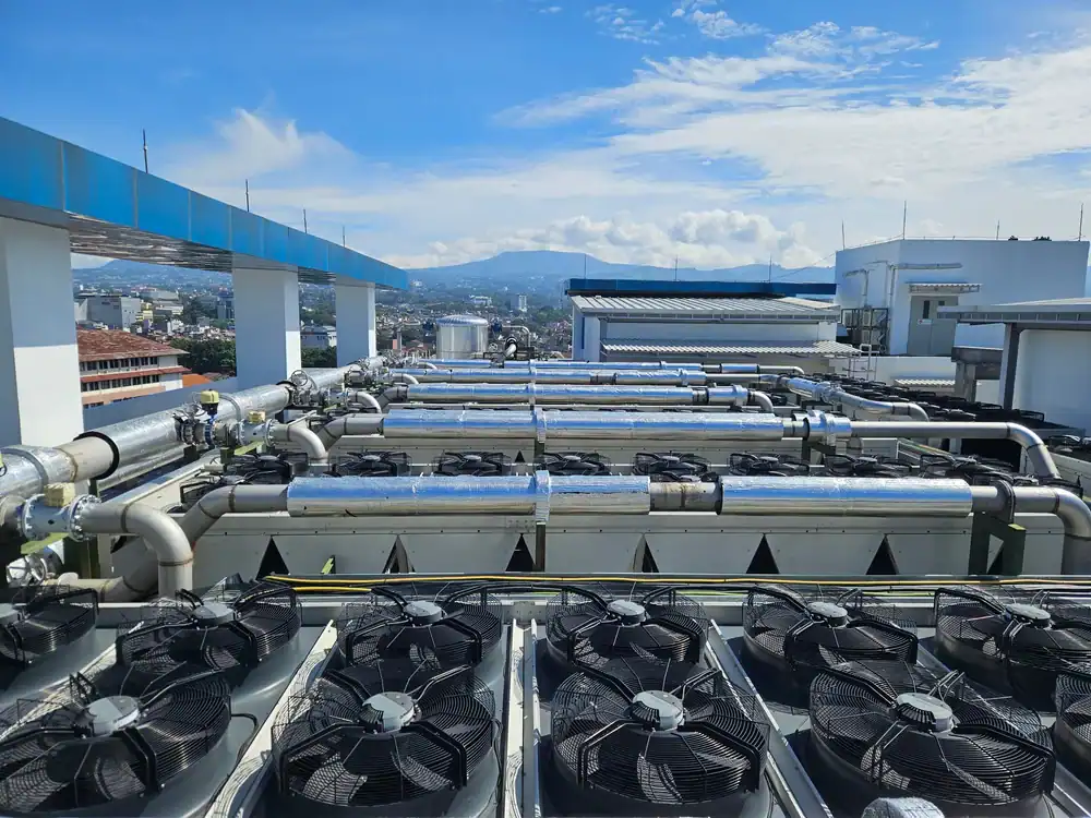 Rows of industrial cooling fans and large pipes on a rooftop under a sunny sky, with a cityscape and mountains visible in the background.