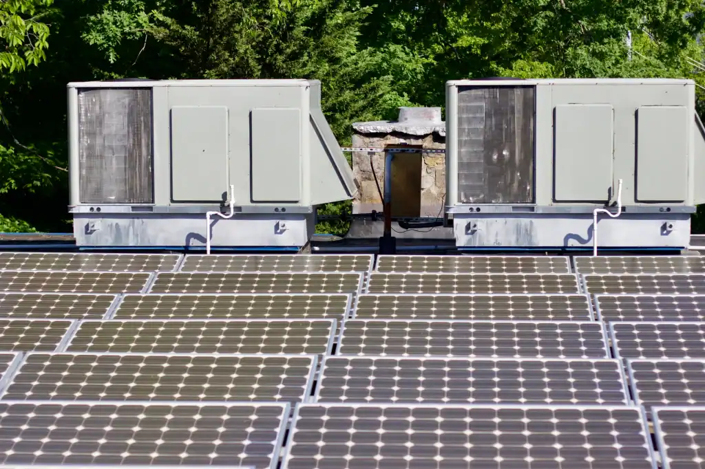Rows of solar panels are installed on a rooftop in front of two large HVAC units, with green trees in the background.