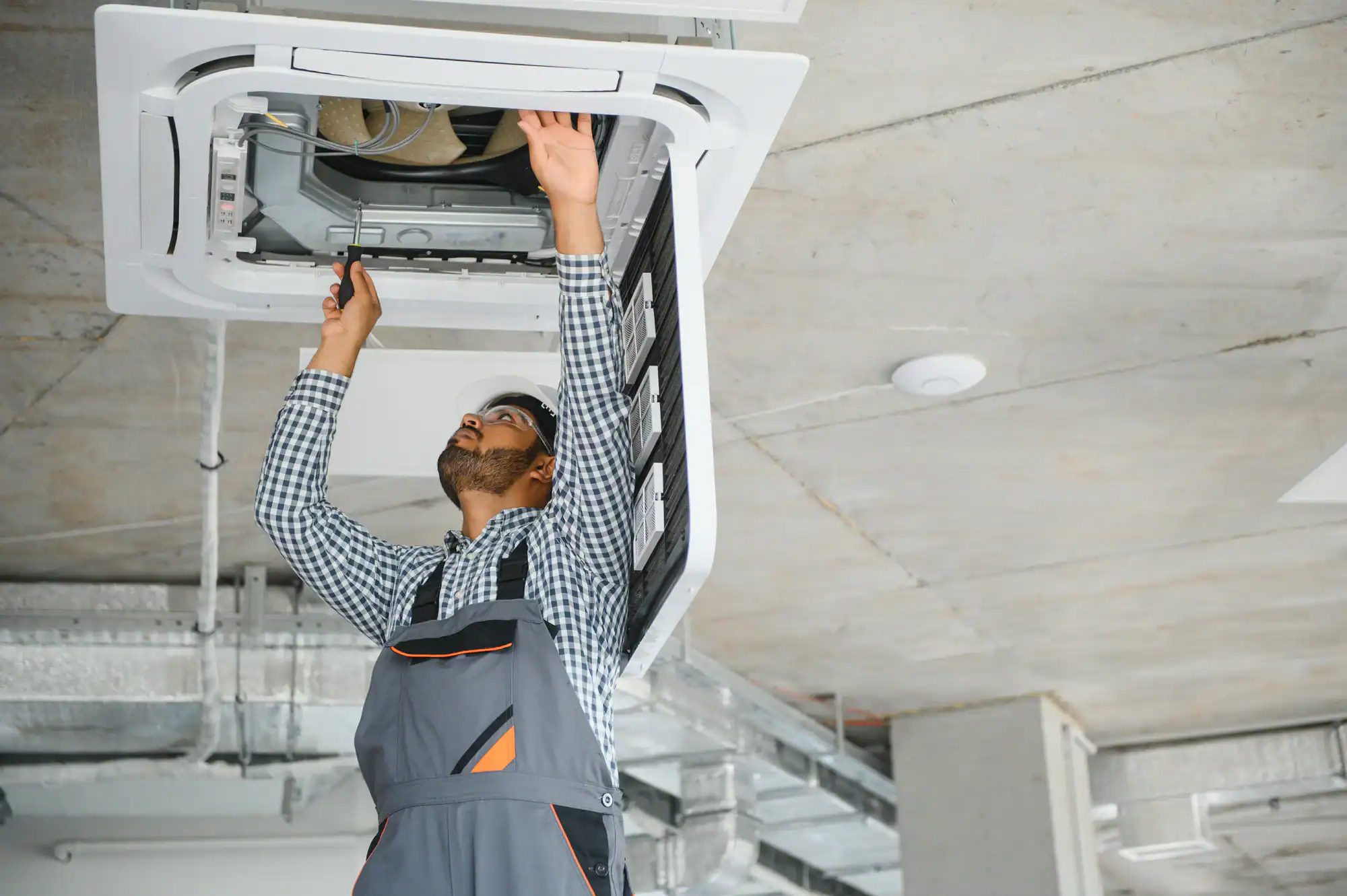 A technician in overalls stands on a ladder and repairs a ceiling-mounted air conditioning unit with a screwdriver in a modern industrial-style room.