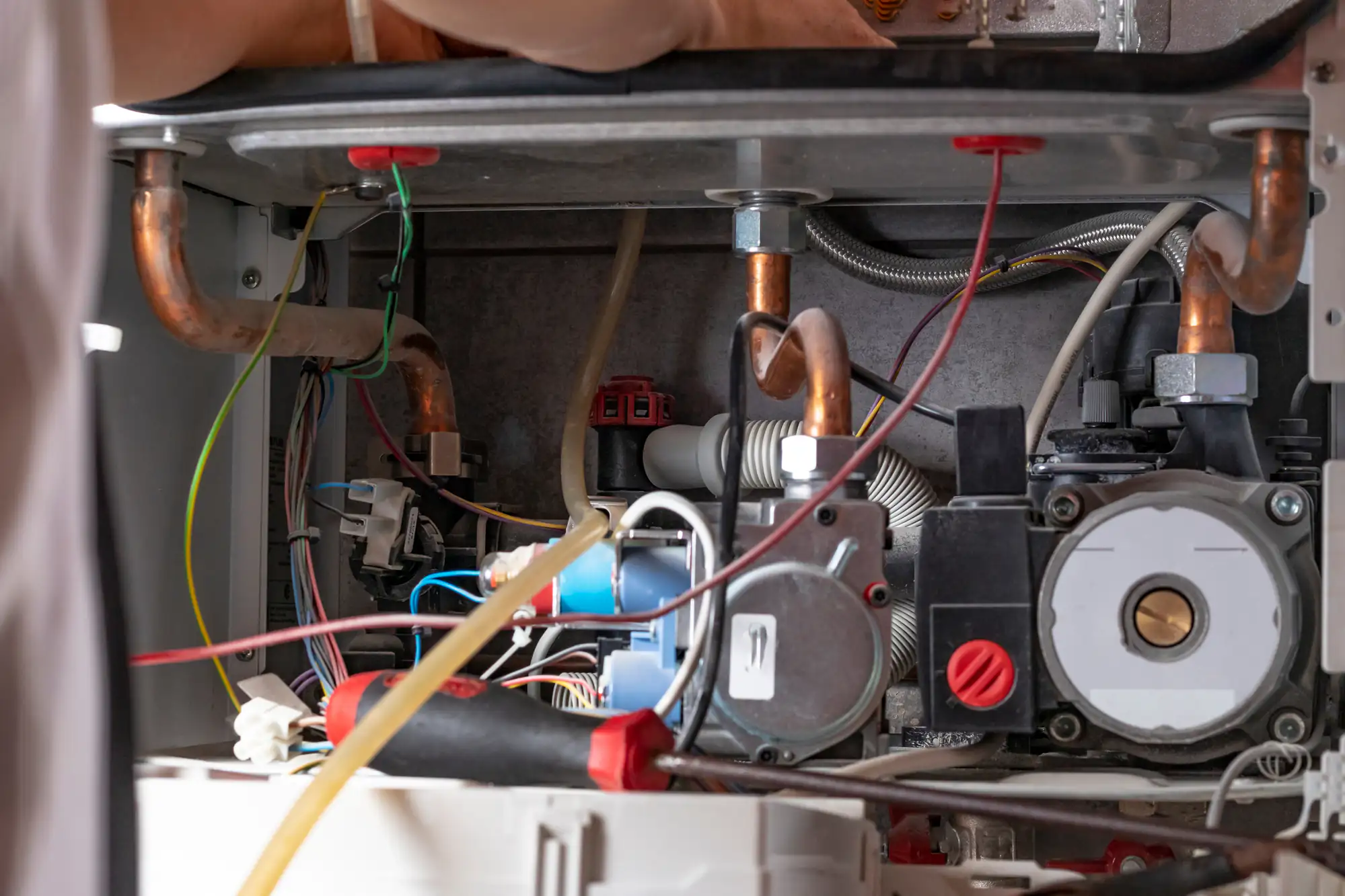 Close-up of a person's hands repairing the internal components of a gas boiler, showing pipes, wires, and various mechanical parts inside the unit.