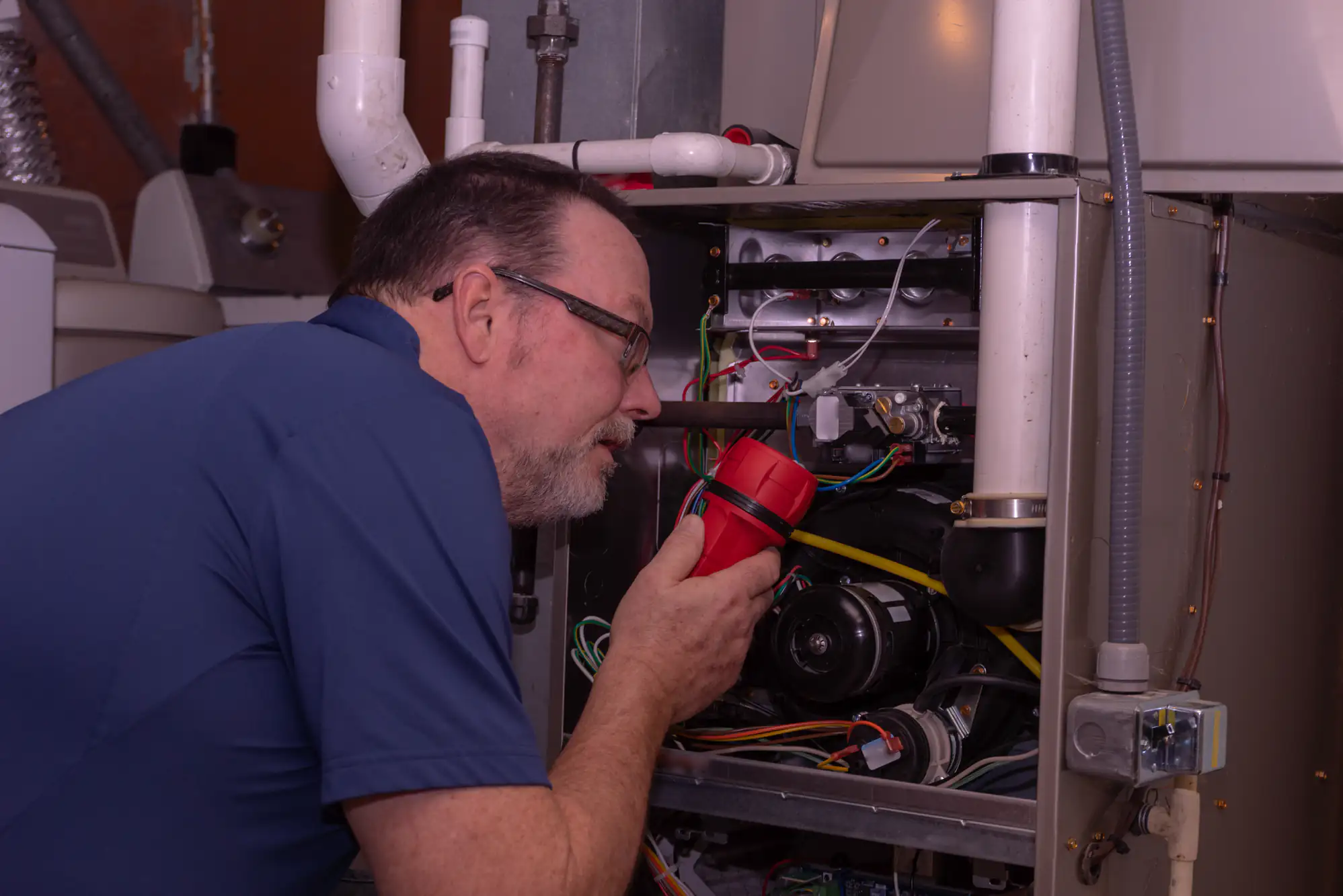 A man wearing glasses examines the inside of a furnace with a red flashlight in a utility room, checking wires and components for maintenance or repair.